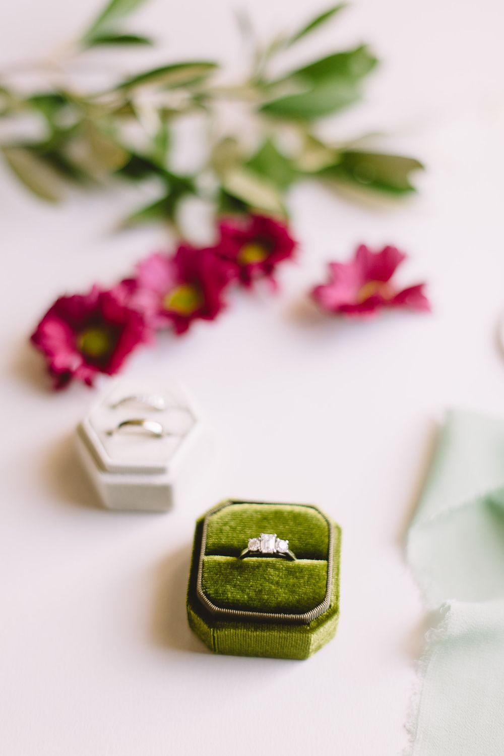 A green velvet ring box with an engagement ring displayed alongside pink flowers and delicate greenery.