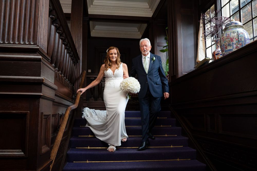Bride in fitted white gown descends grand staircase with father on blue carpeted steps.