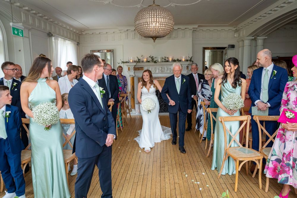 Bride walks down aisle with father past sage green bridesmaids in bright modern ceremony room.
