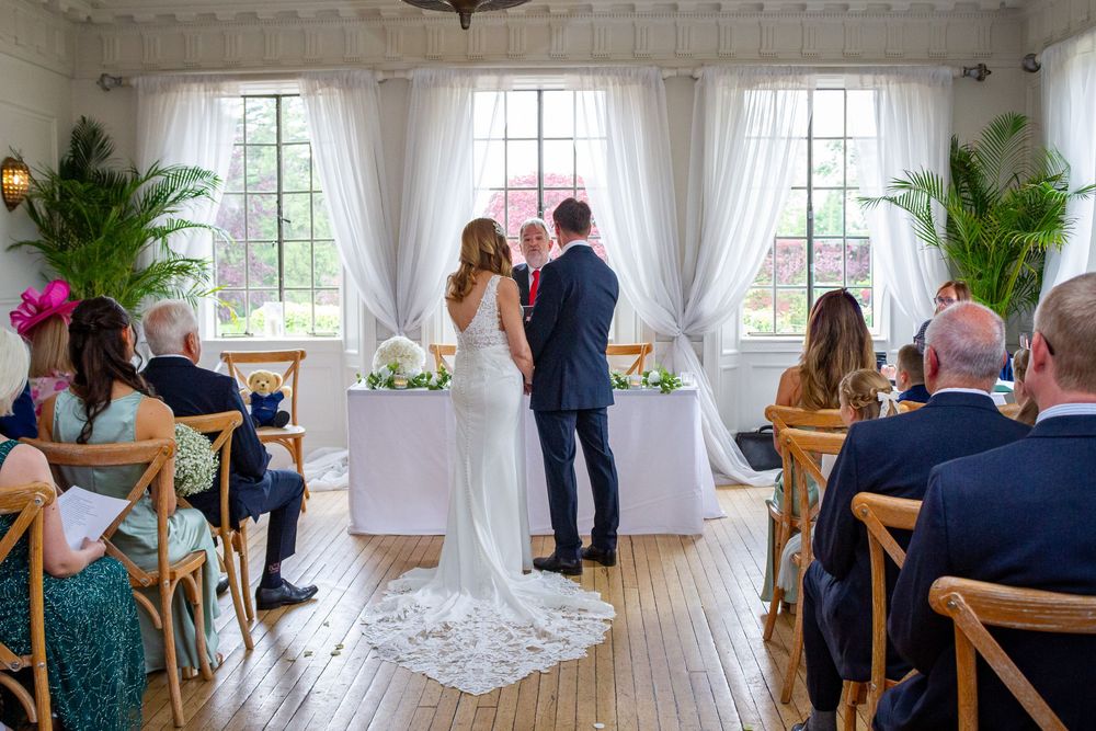 Bride and groom exchange vows at altar in sunlit white-draped ceremony room with tropical plants.