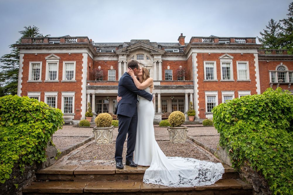 Bride and groom share a kiss on steps outside a grand red-brick English country house wedding venue.