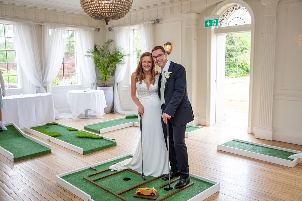 Smiling bride and groom pose together beside a mini golf course set up inside a bright wedding venue.