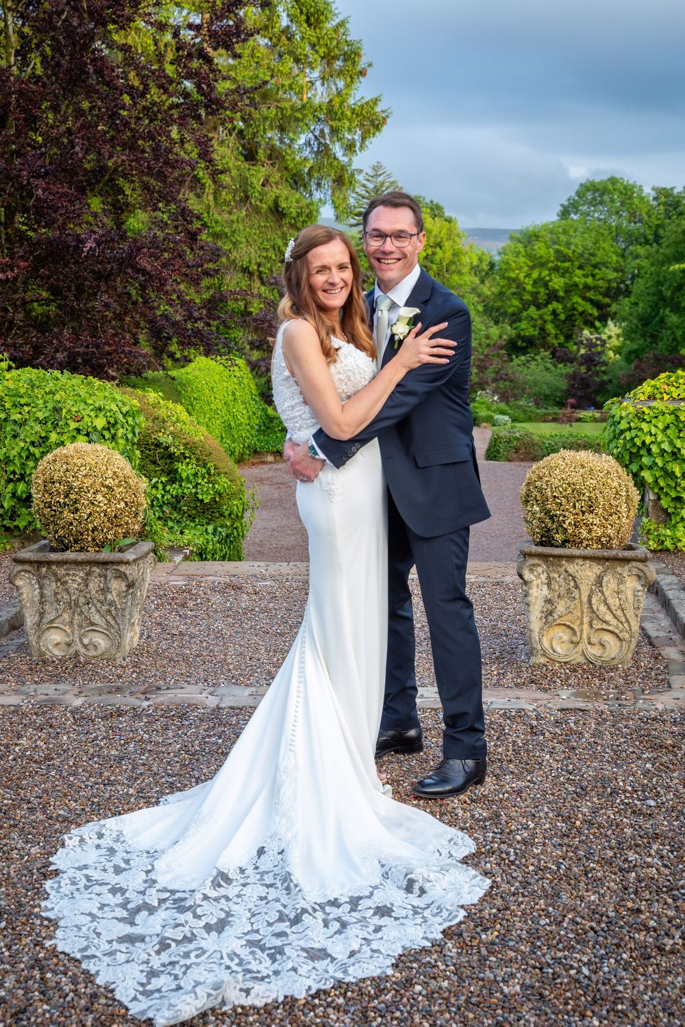 Happy bride and groom embrace in a beautiful formal garden, brides lace train spread elegantly on gravel.