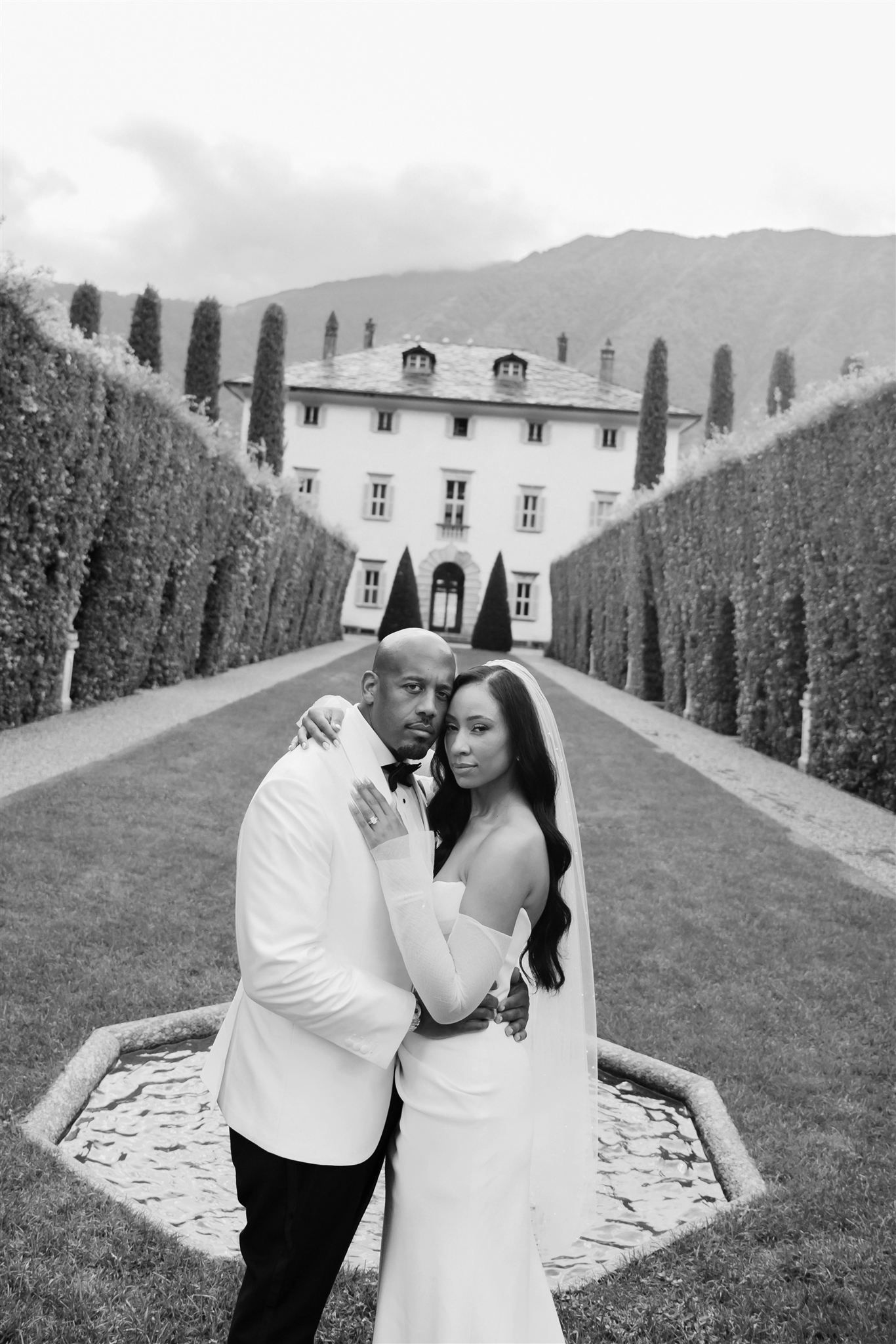 Couple in wedding attire sharing intimate moment in formal garden with mansion backdrop.
