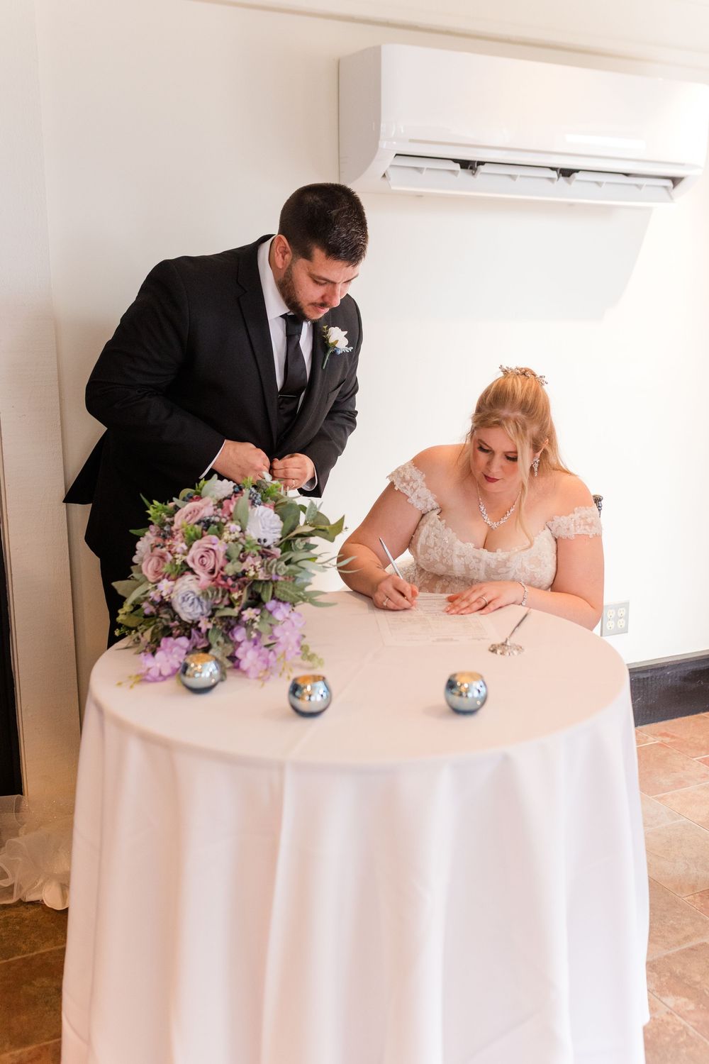 Couple signing marriage documents at a round table with floral centerpiece in elegant wedding ceremony setting.