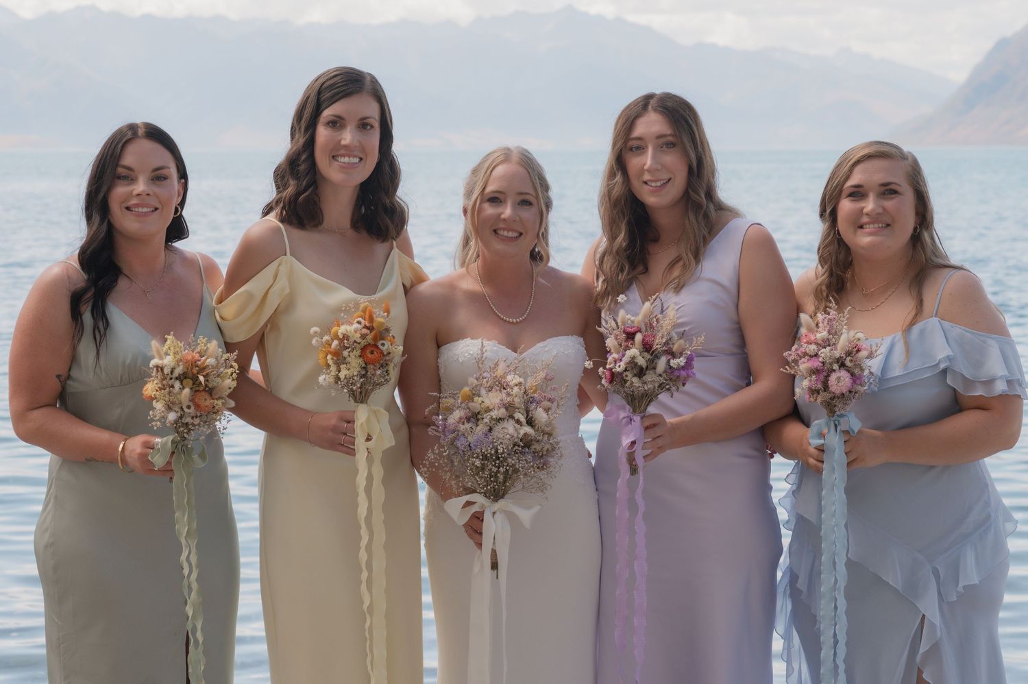 A group of bridesmaids in pastel dresses stand together holding bouquets on a beach with mountains in the background.