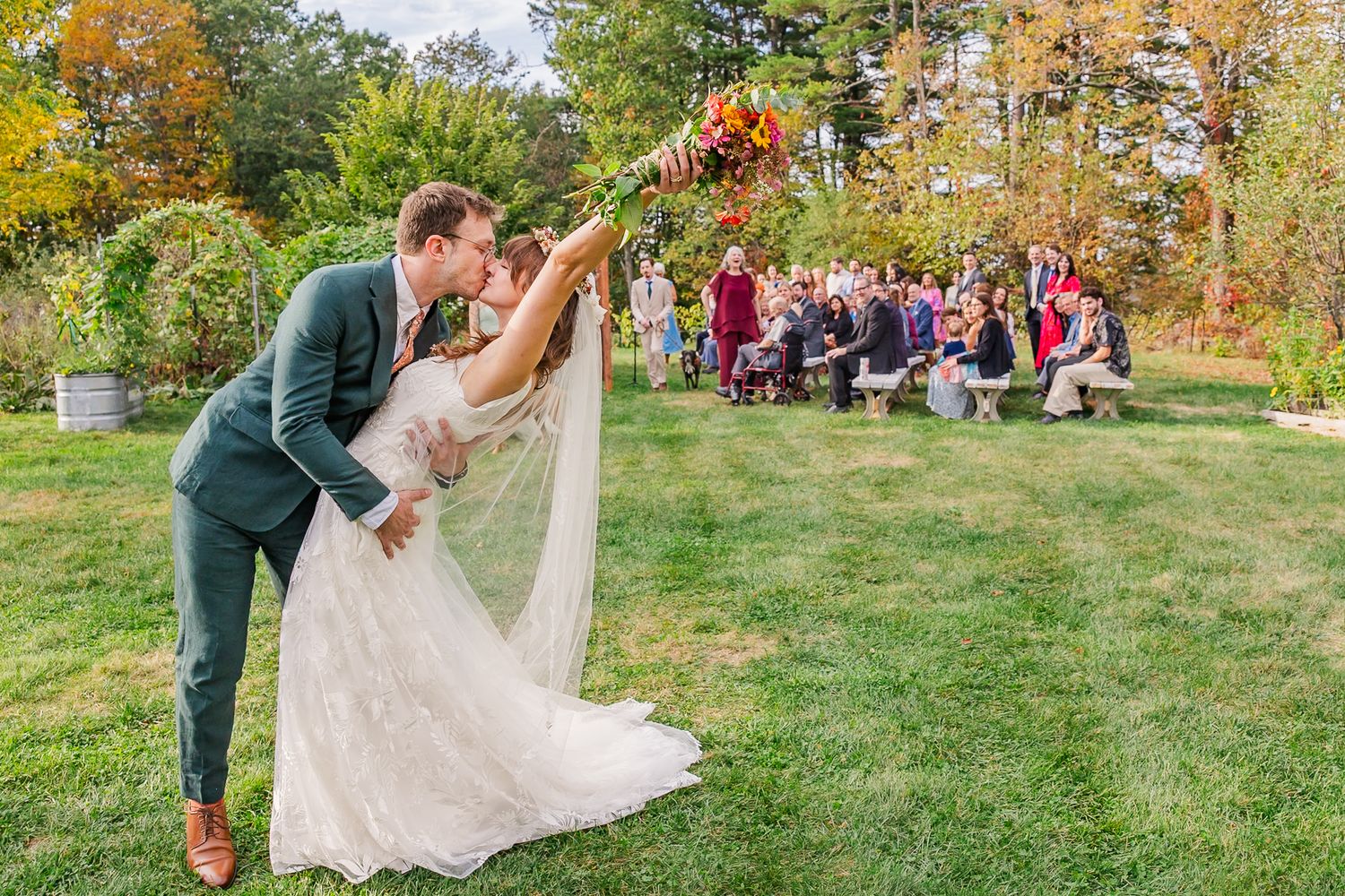 A couple shares a romantic moment during an outdoor autumn wedding ceremony with guests seated on wooden benches.