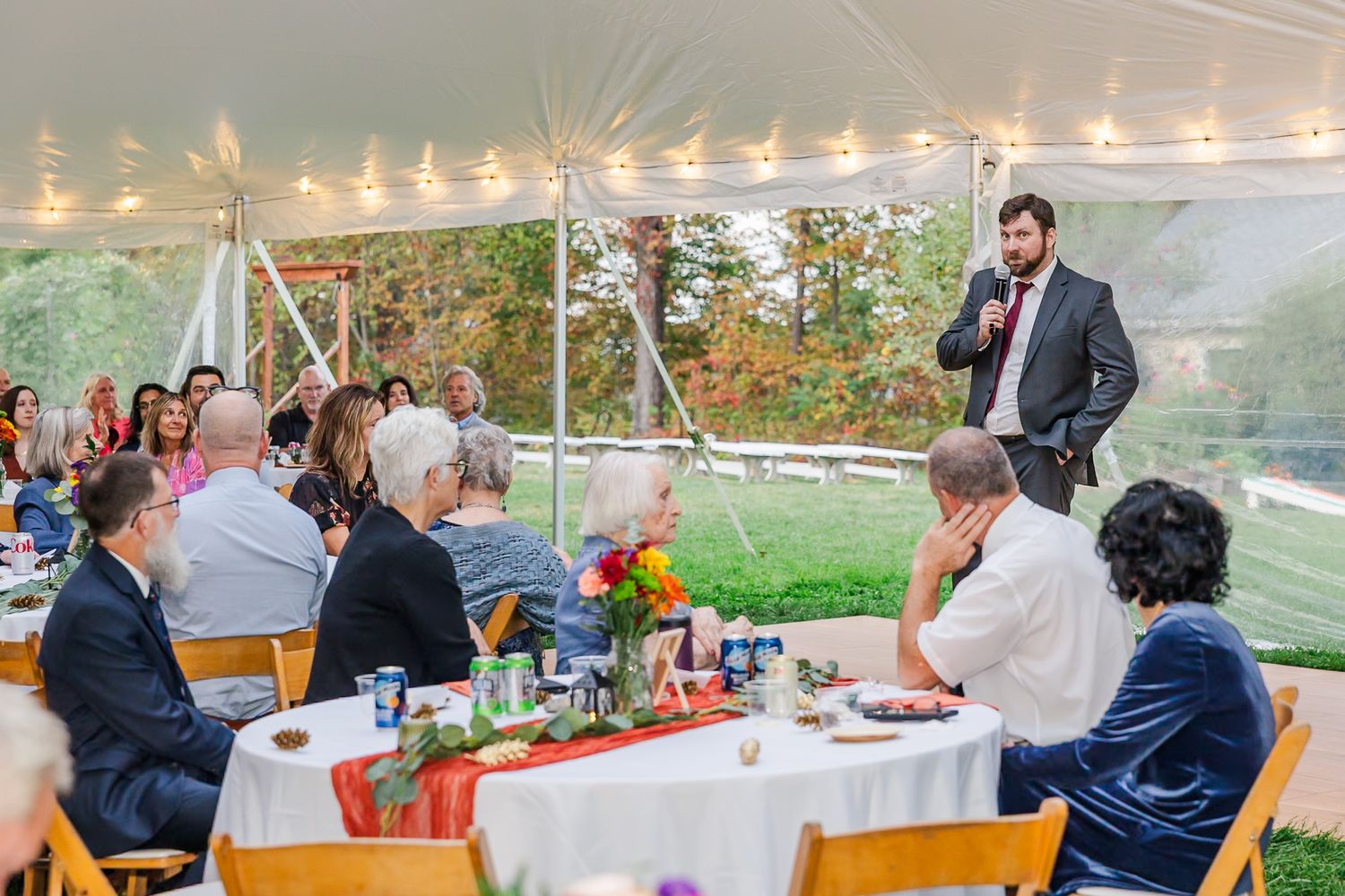 Wedding guests seated at tables under an outdoor tent while someone gives a speech during an autumn reception celebration.