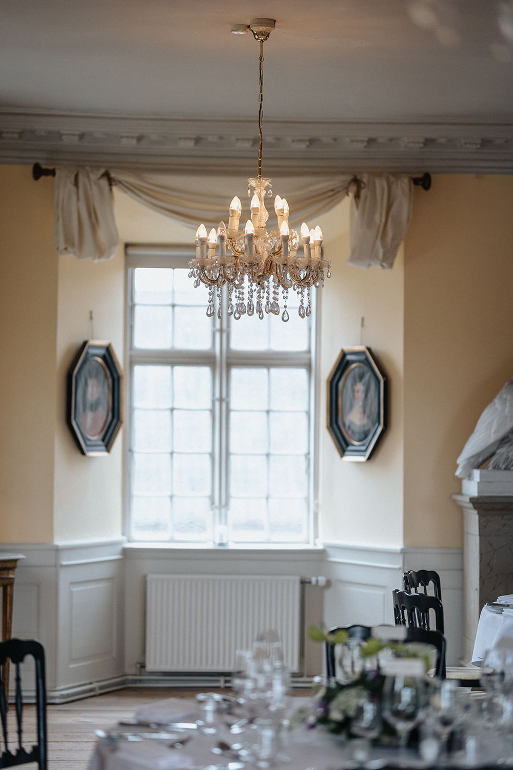 Crystal chandeliers hang elegantly from the ceiling in a classic dining room with white wainscoting and hexagonal mirrors.
