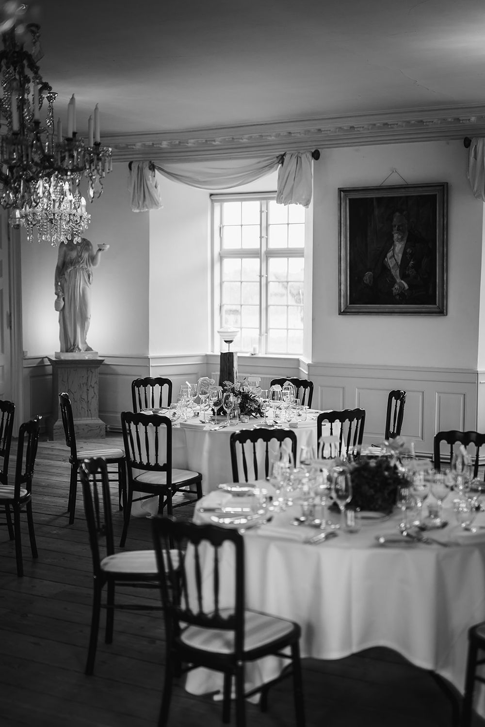 Elegant black and white photo of a formal dining room with round tables, crystal chandelier and classic artwork on walls.
