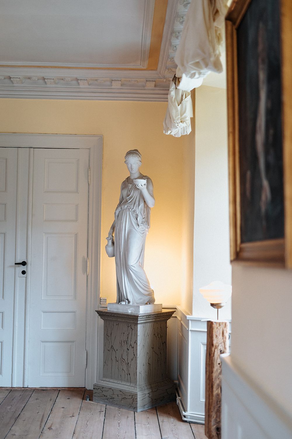Classical white marble statue on decorative pedestal stands elegantly in corner of historic room with yellow walls.