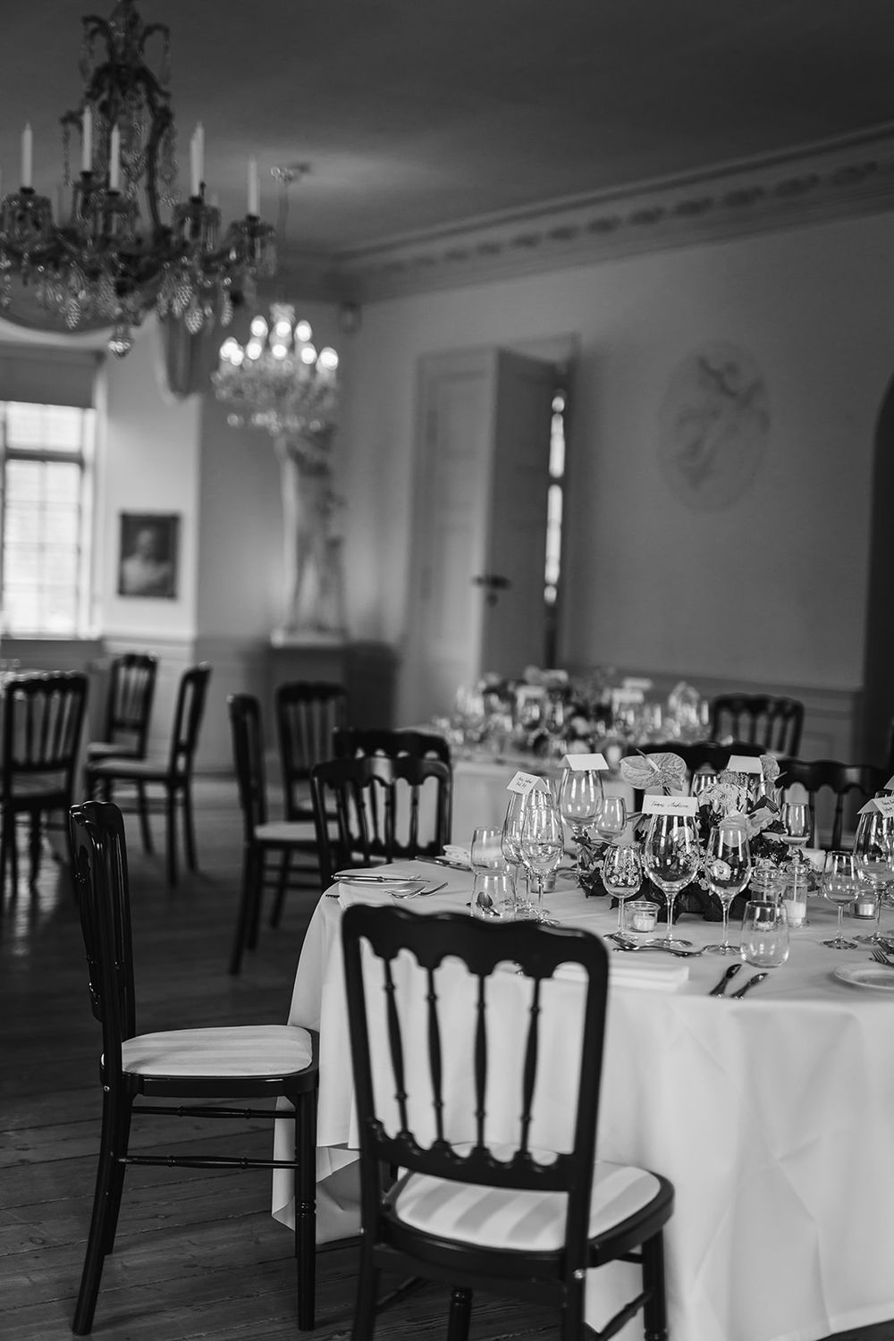 Black and white elegant banquet hall setup with round tables, white tablecloths, and dark wooden chairs under chandeliers.