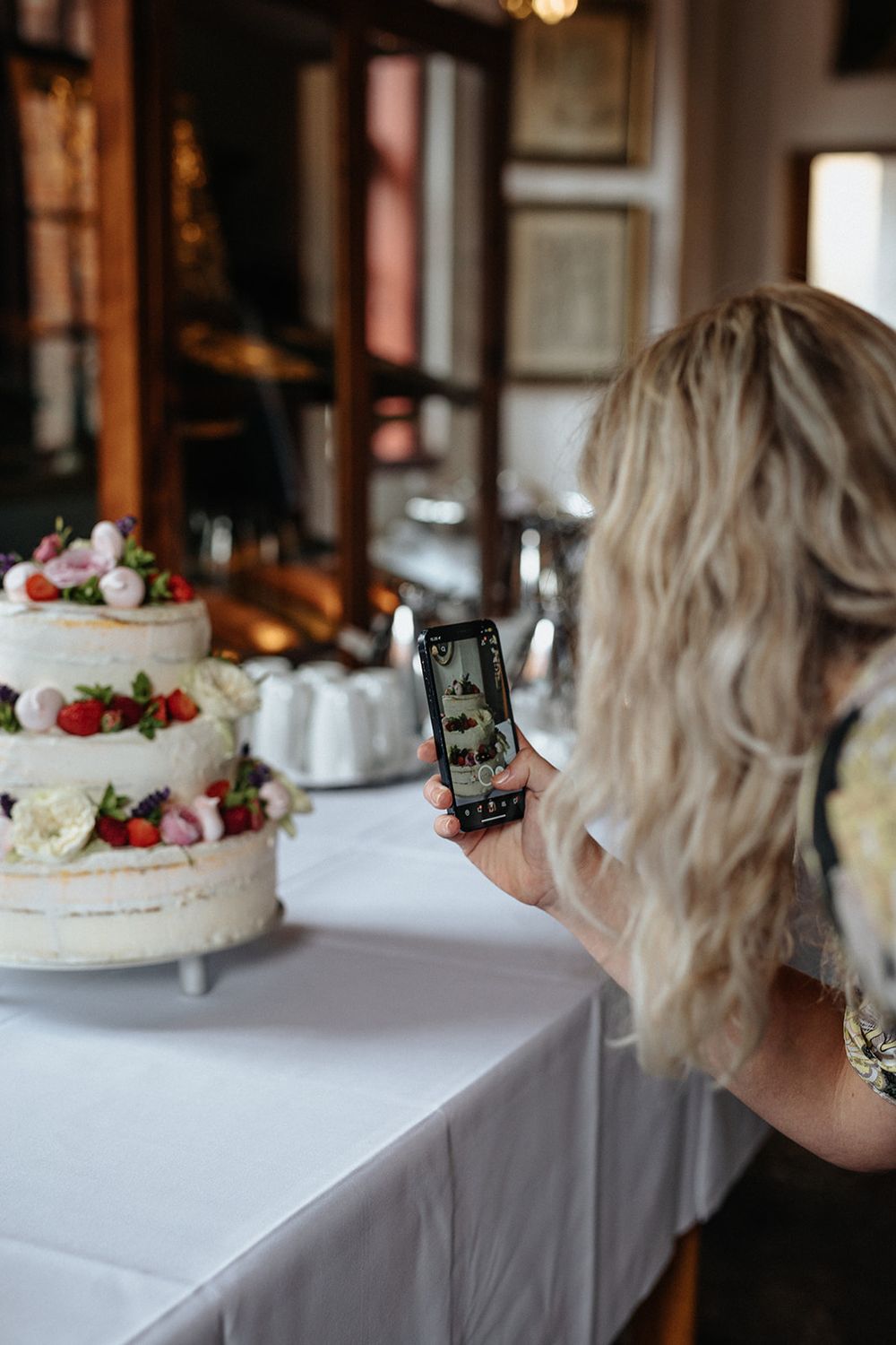Someone taking photos of an elegant white wedding cake decorated with fresh berries and flowers on a table.