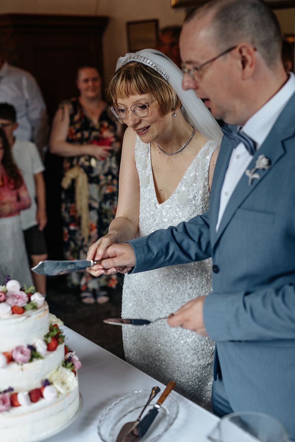 A couple cuts their wedding cake together, with the bride wearing a white dress and veil and groom in a blue suit.