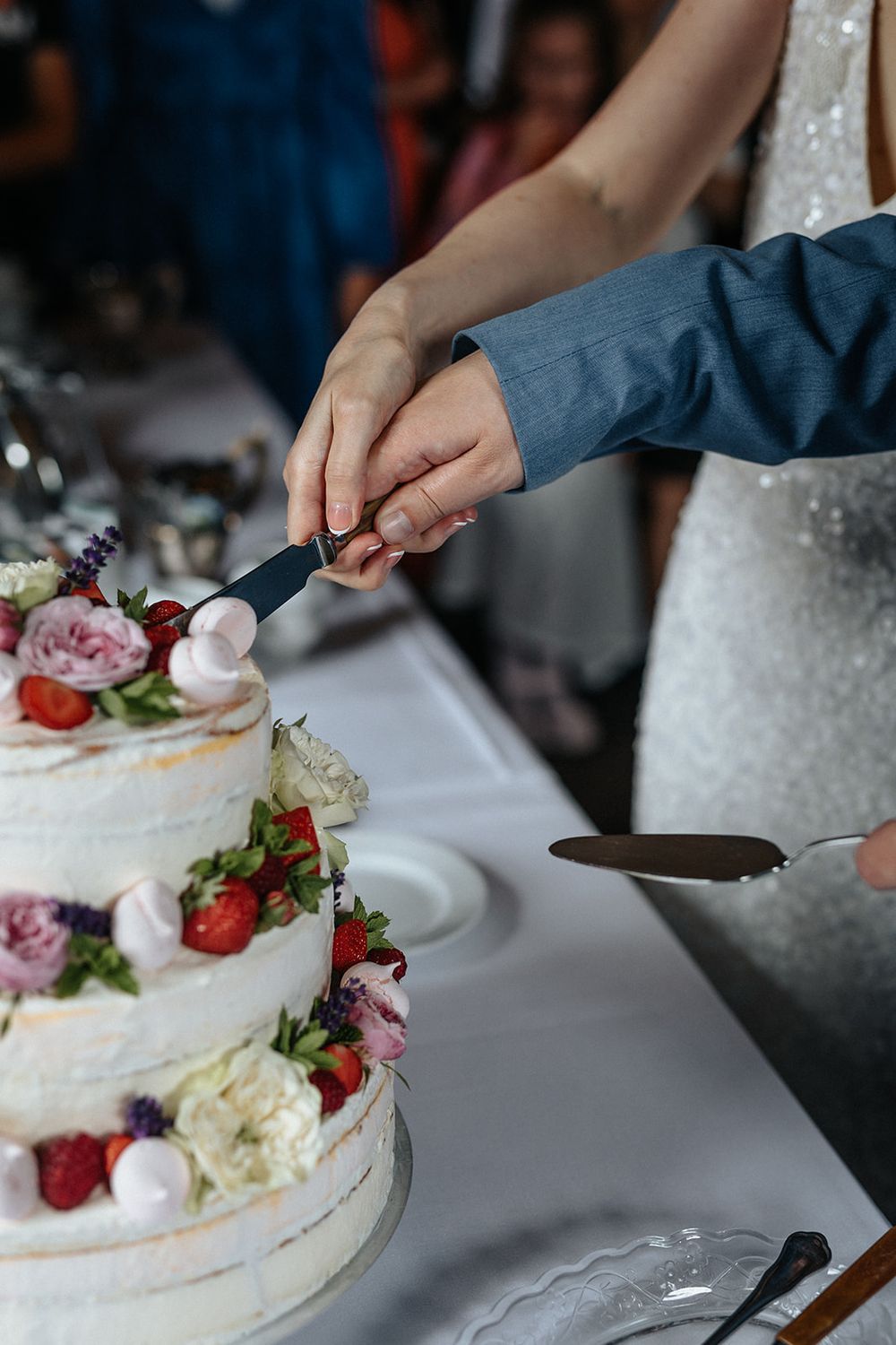 Couple's hands cutting into a beautiful tiered wedding cake decorated with fresh flowers and strawberries.