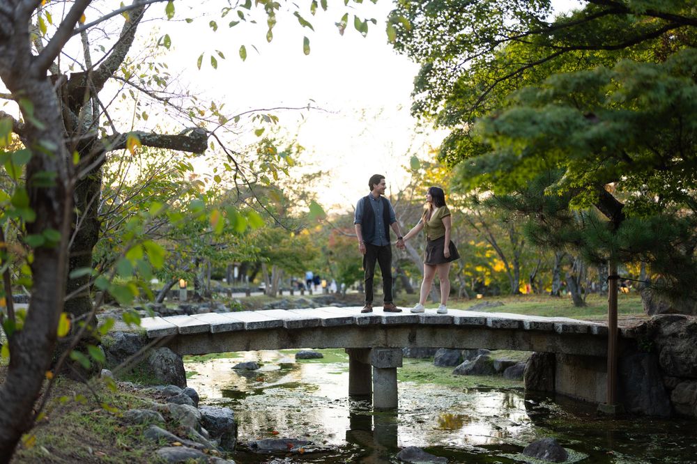 Couple standing together on a stone arch bridge surrounded by green trees in a serene Japanese park.