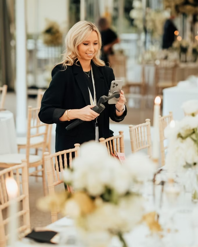 A woman in a black blazer sits at an elegant wedding reception table taking photos of the floral centerpieces.