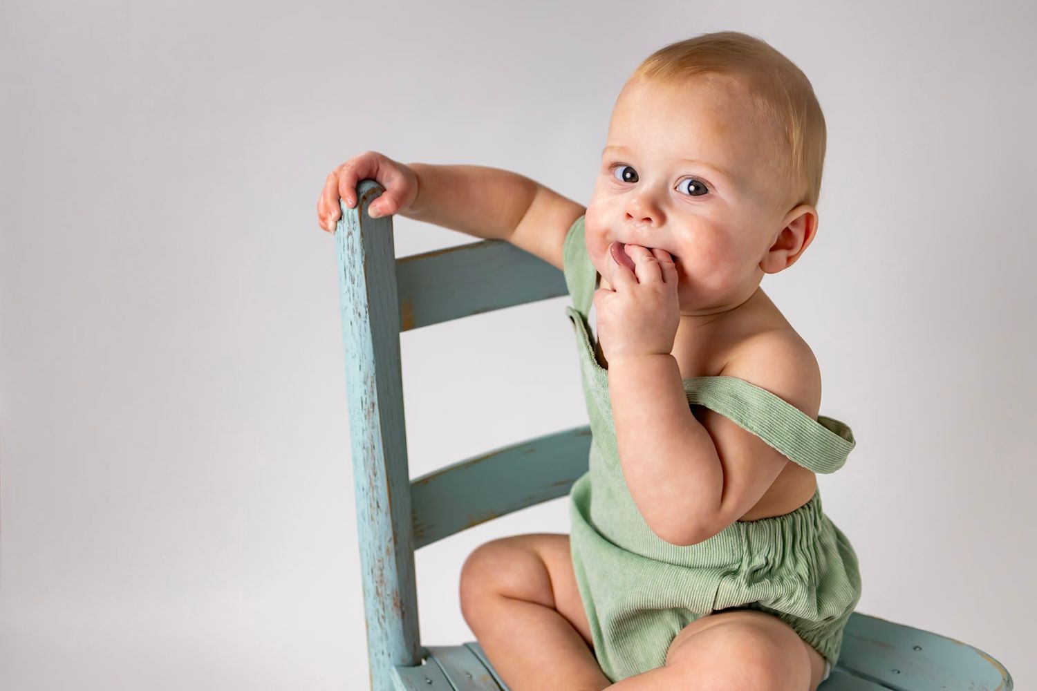 A small child in a mint green romper sits and plays on a wooden chair in a series of candid poses.