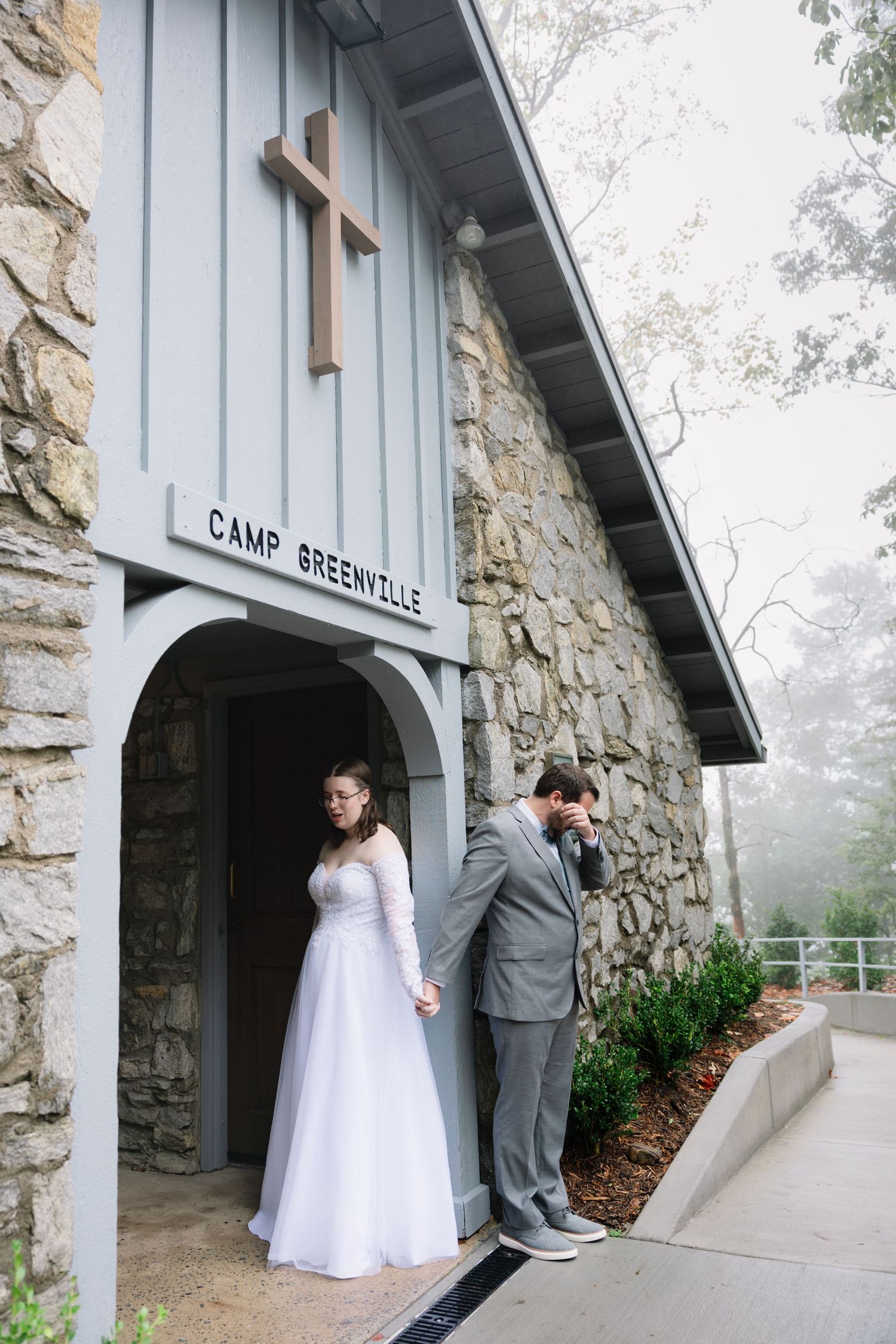 A wedding couple shares a tender moment outside a rustic stone building with 'Camp Greene' signage in misty weather.