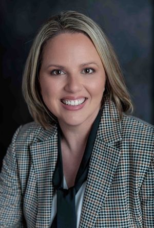 Smiling professional woman with blonde hair in houndstooth blazer, business portrait dark background.