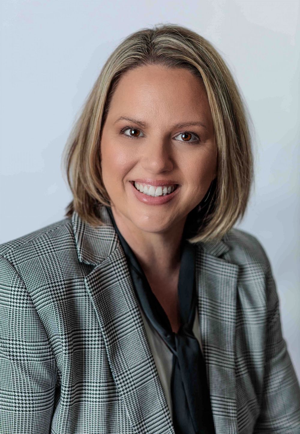 Professional blonde woman smiling in houndstooth blazer, business headshot on white background.