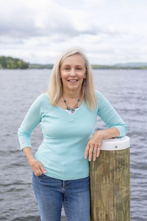 Smiling blonde woman in teal top leaning on a dock post with lake in background.