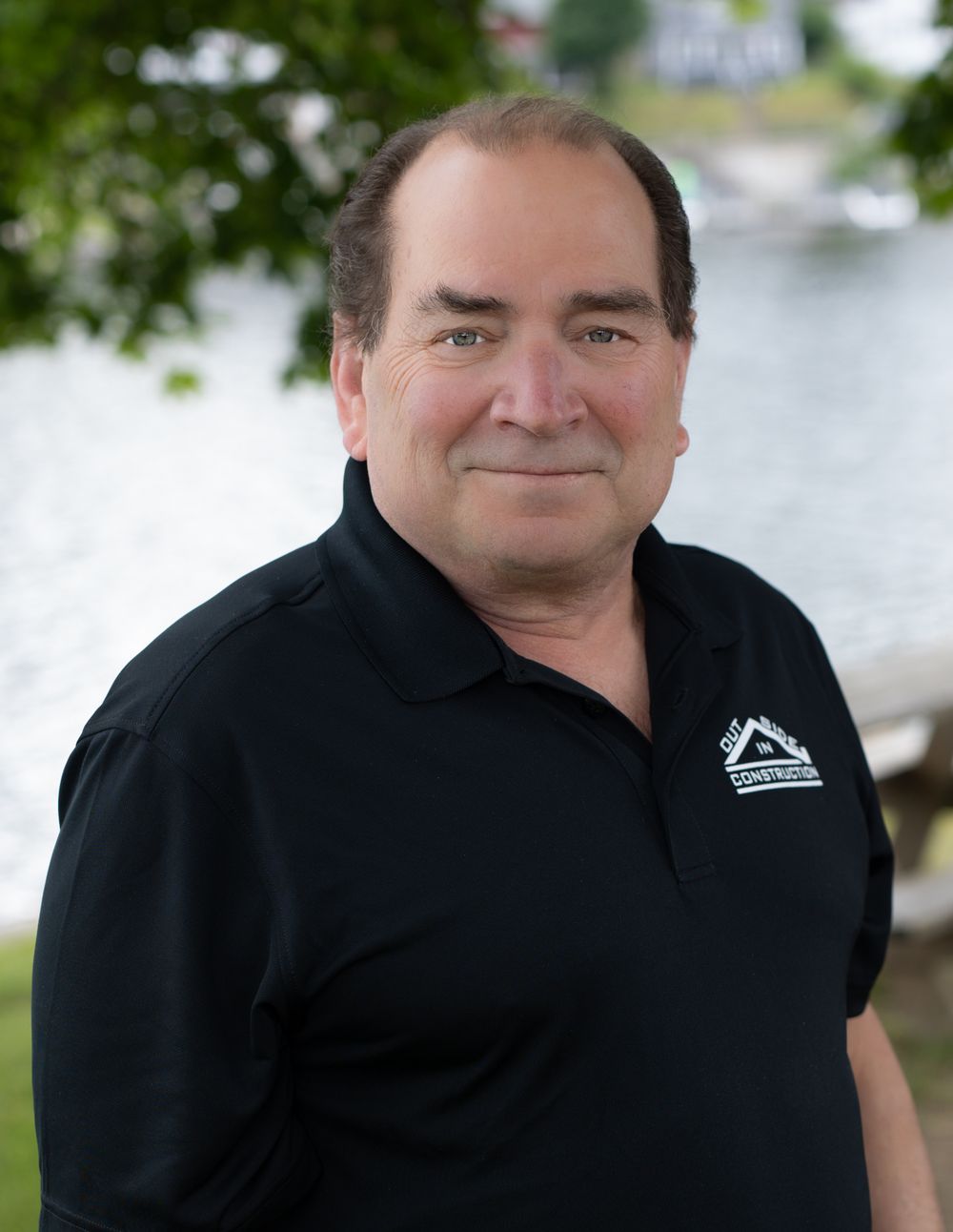 Middle-aged man smiling outdoors in black polo shirt with company logo, water and trees in background.