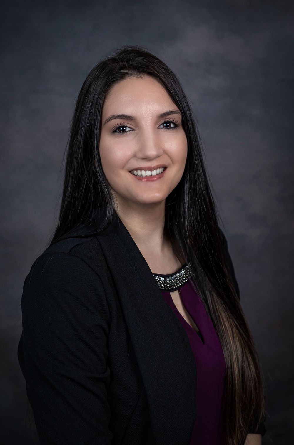Young woman with long dark hair smiling in black blazer and purple top, professional headshot.