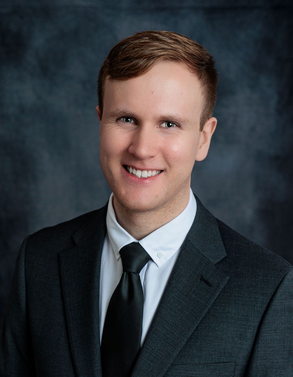 Young professional man in gray suit and dark tie smiling, formal business portrait headshot.