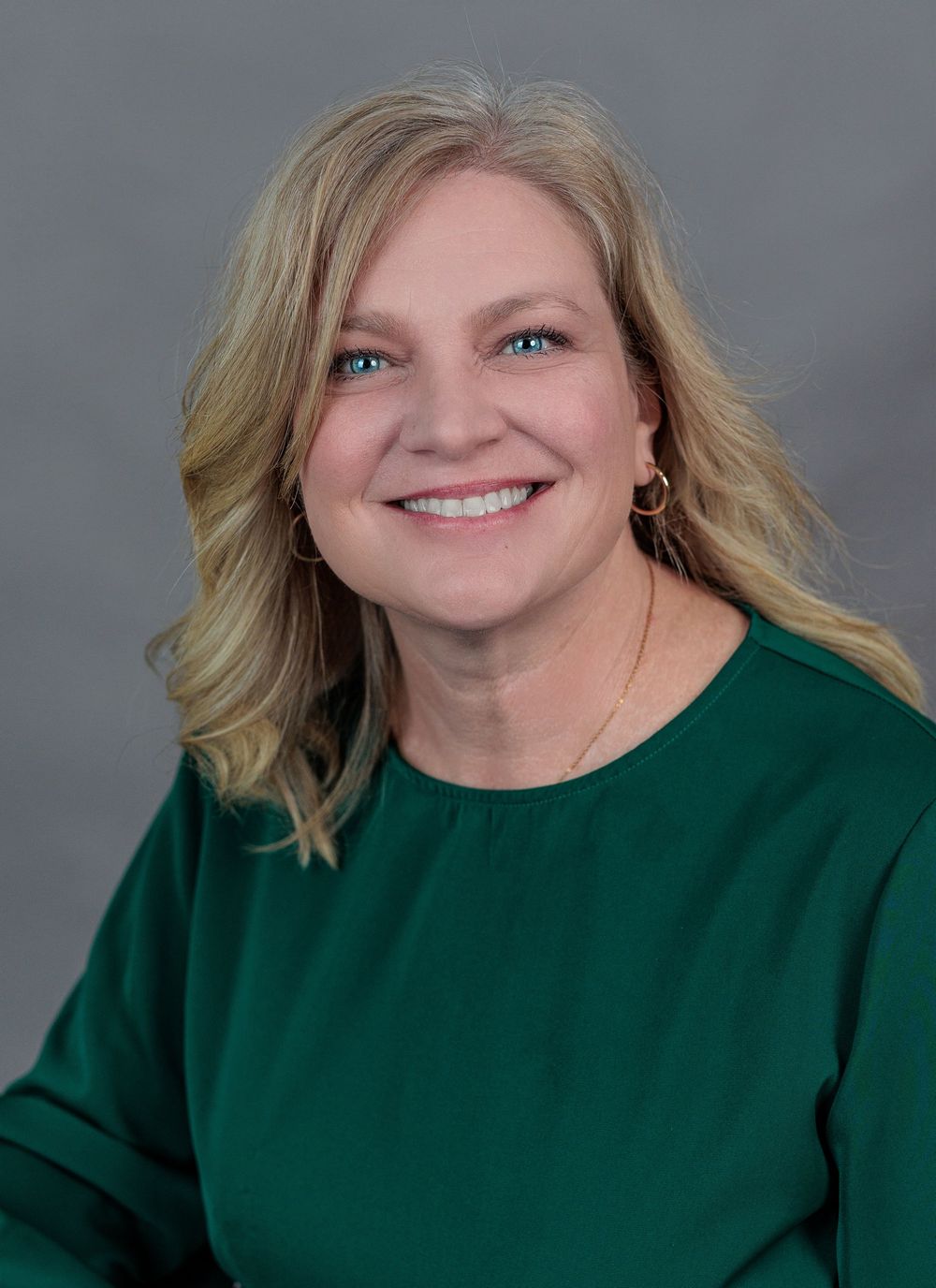 Smiling middle-aged blonde woman in green top with gold hoop earrings, professional headshot.