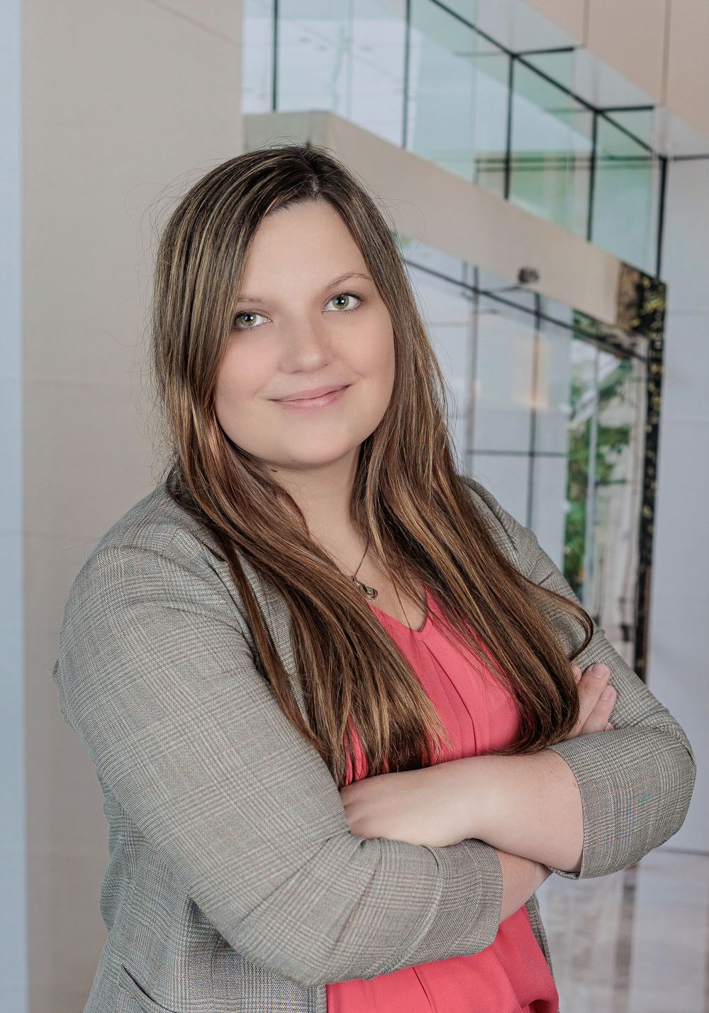Young woman with long brown hair smiling in gray blazer, arms crossed, modern interior background.