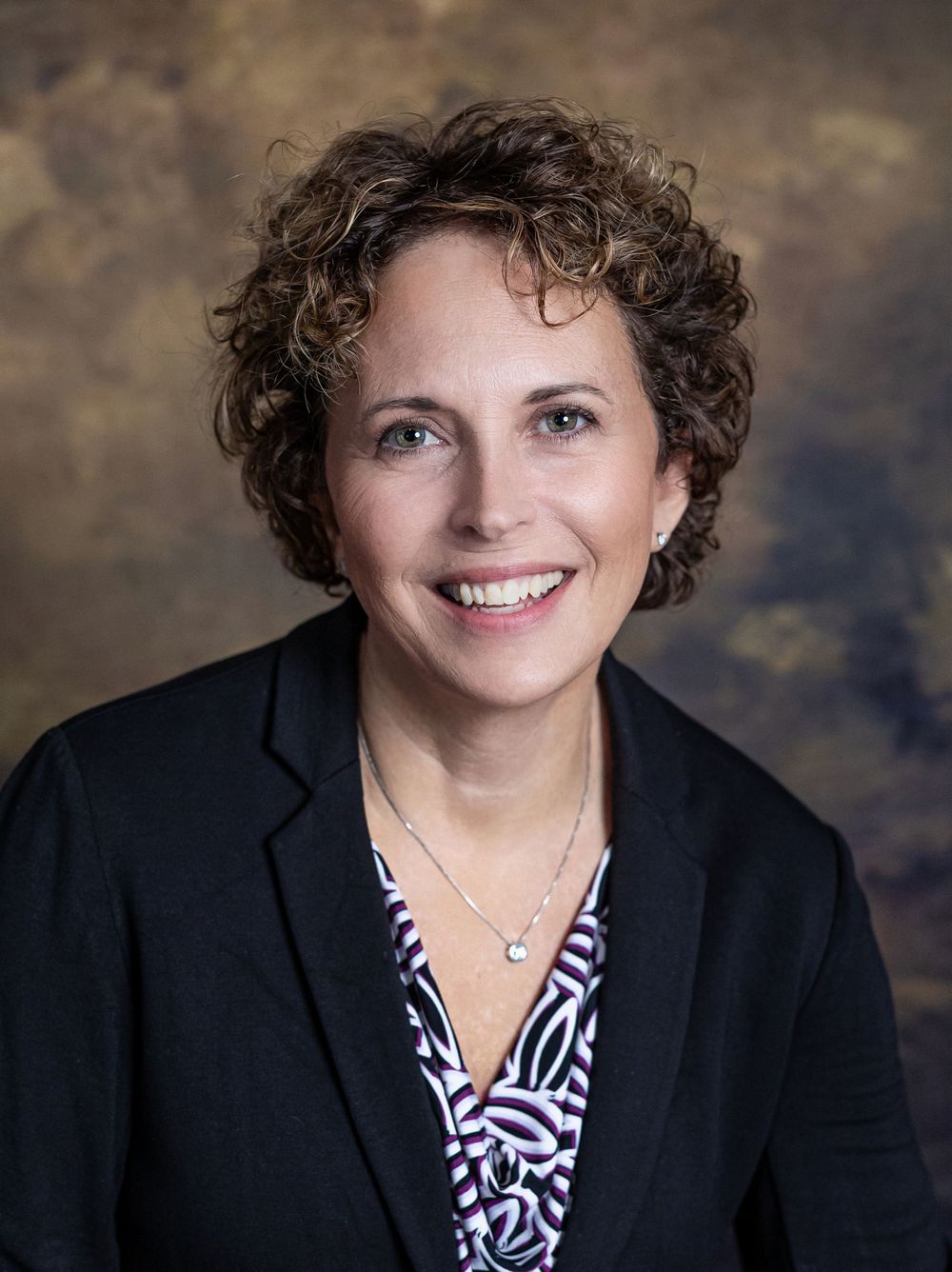 Professional woman with curly brown hair in black blazer and patterned blouse, business portrait.