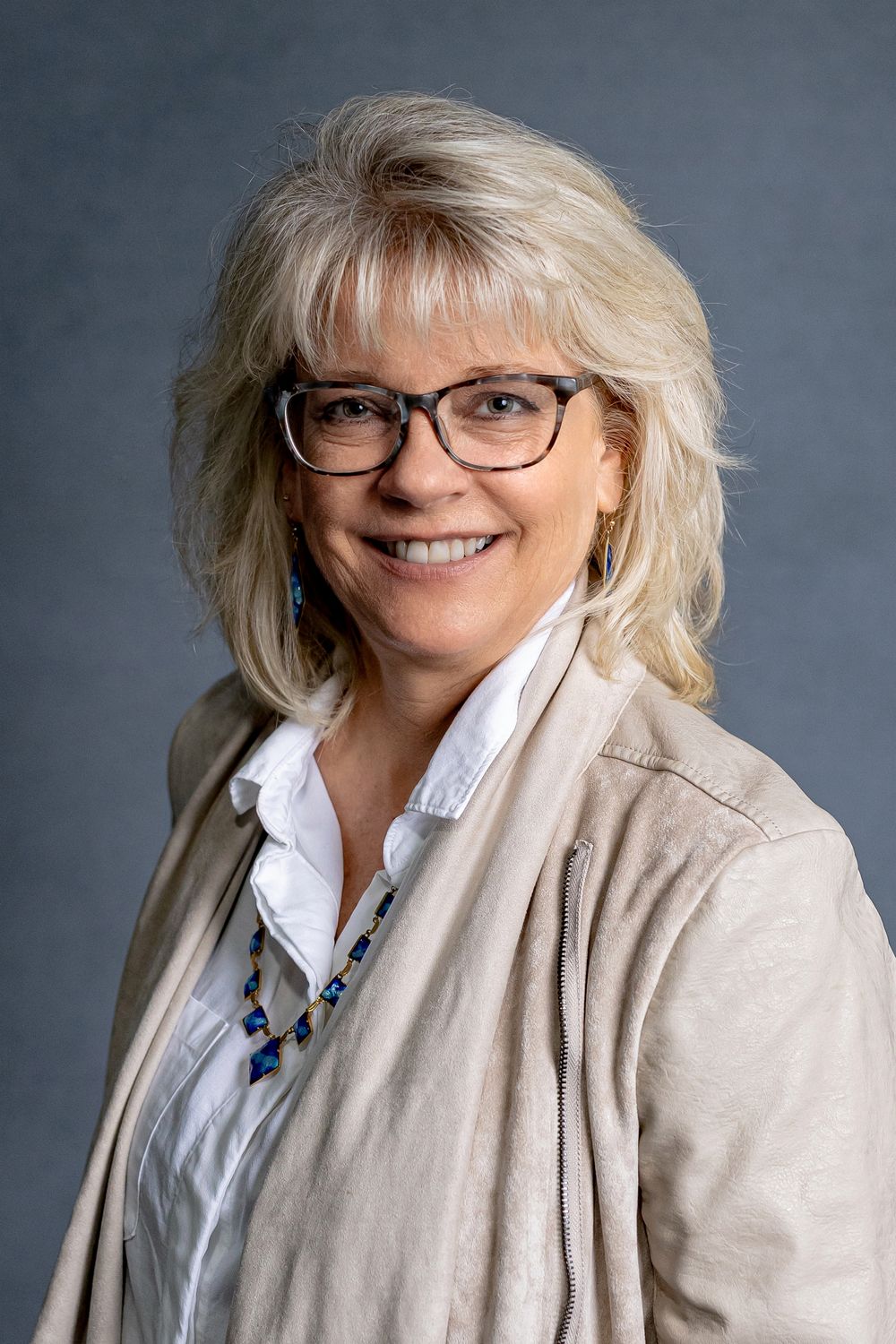 Smiling woman with blonde hair and glasses in beige jacket with blue necklace, professional photo.