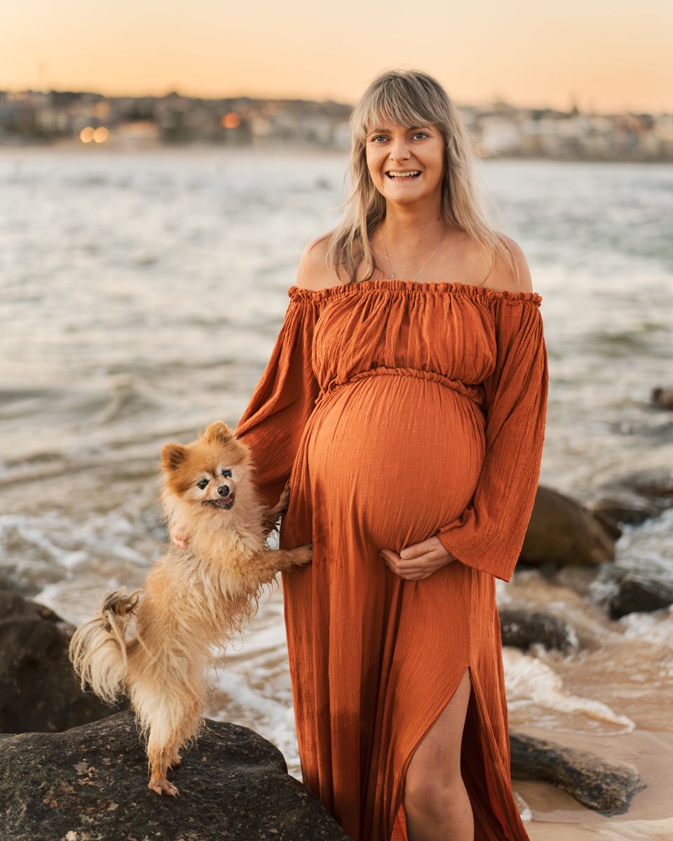 Pregnant woman stands along a sandy beach at sunset with her cute small dog during a maternity photoshoot in Sydney