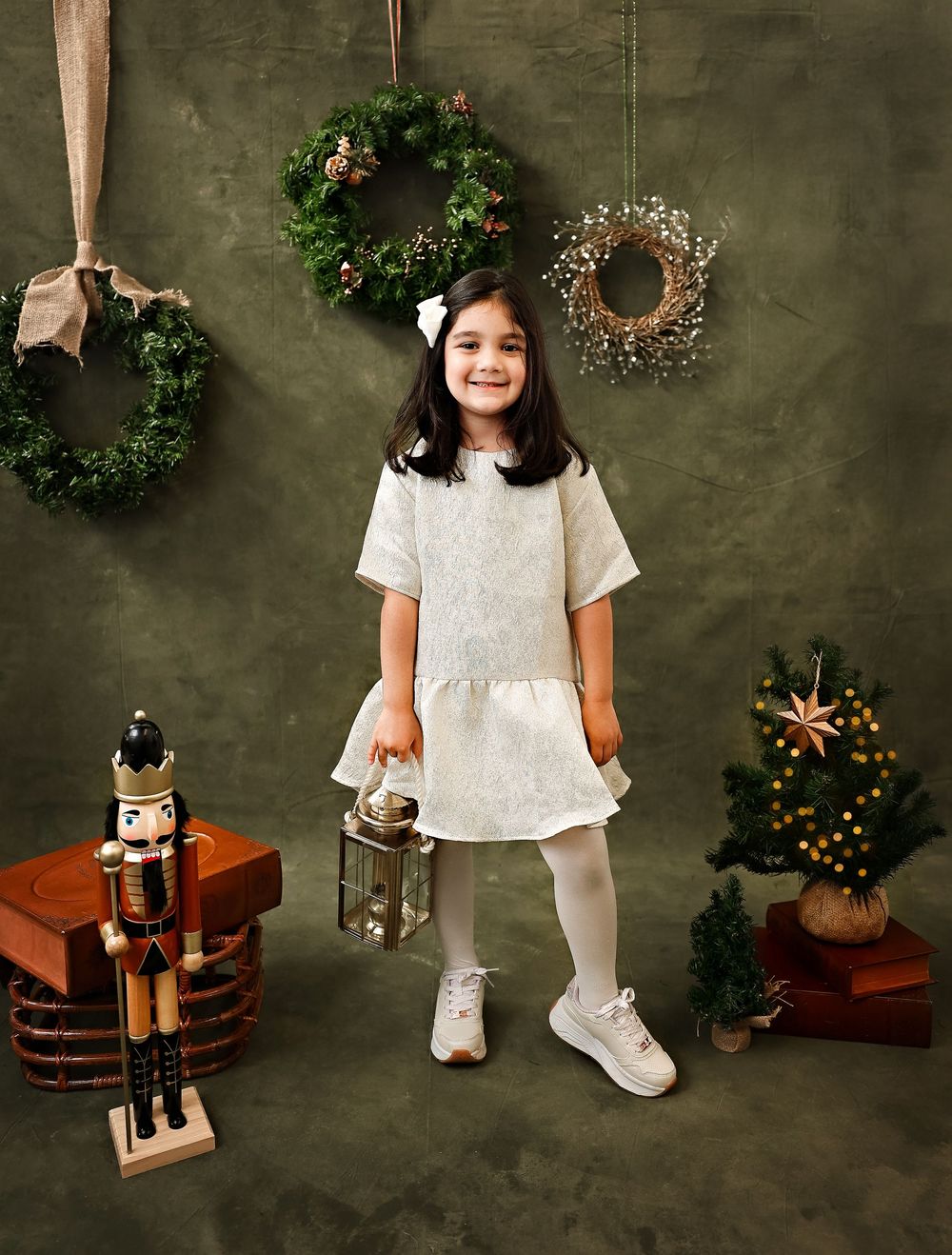 Holiday portrait with white dress, decorative wreaths, nutcracker, and lantern against green backdrop with Christmas decor.