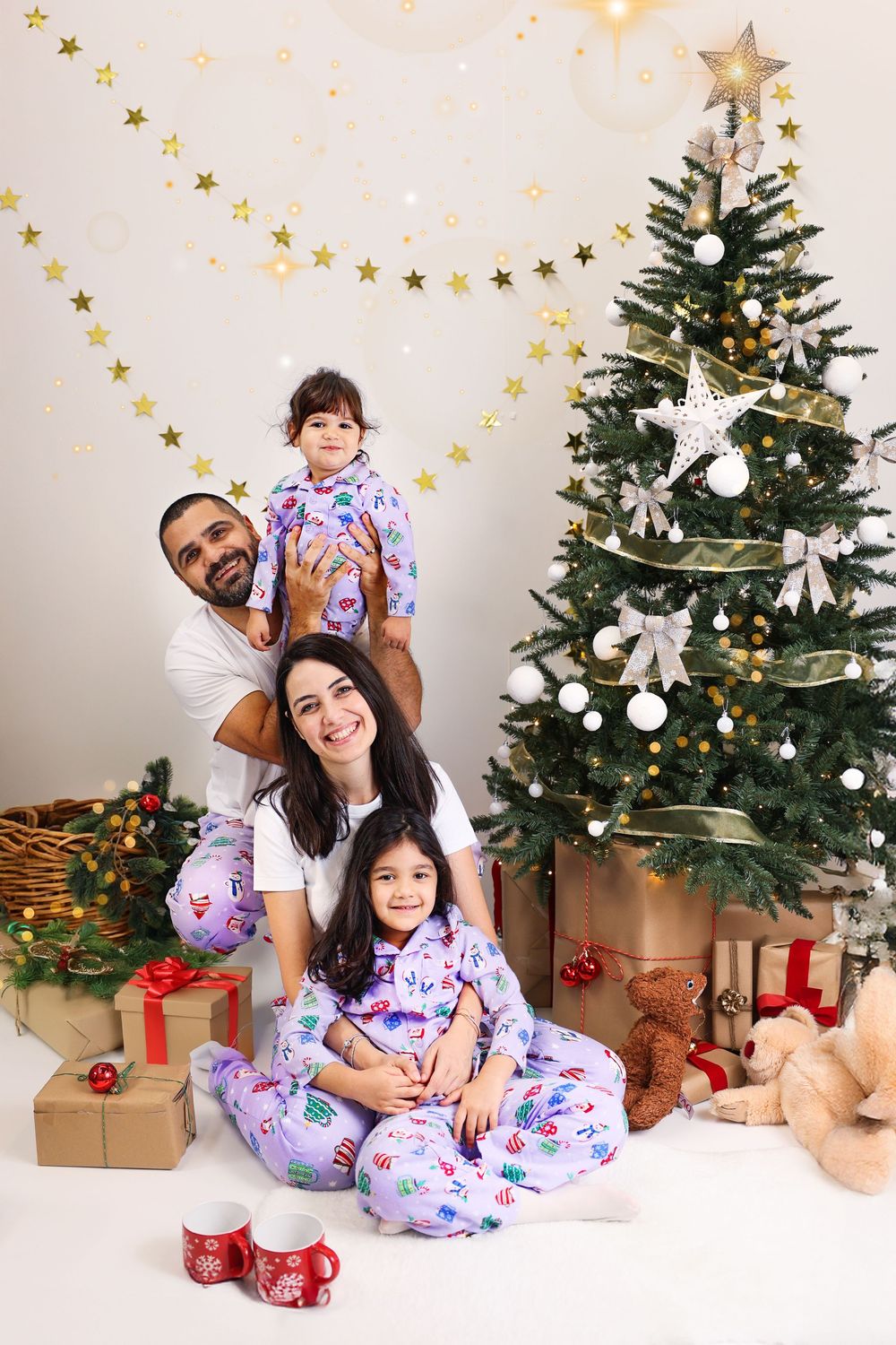 A decorated Christmas tree stands beside wrapped presents and gold star garlands against a white wall.