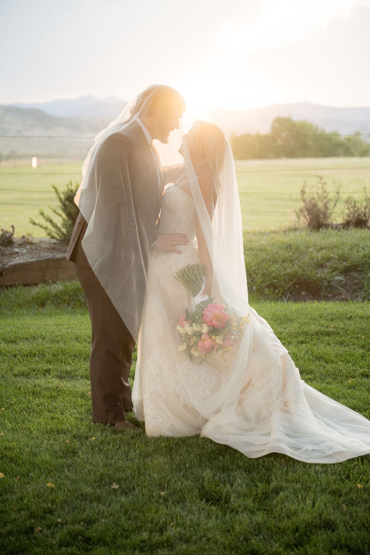 A romantic wedding couple embraces in a sunlit meadow with mountains in the background as the bride's veil flows in the breeze.