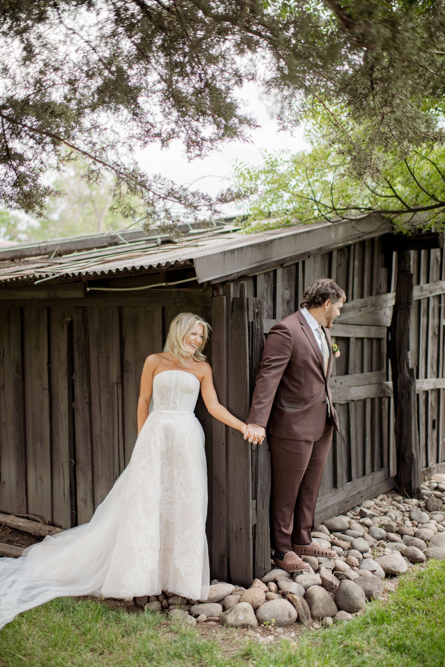 A couple in wedding attire shares a tender moment behind a rustic wooden fence with stone foundation.