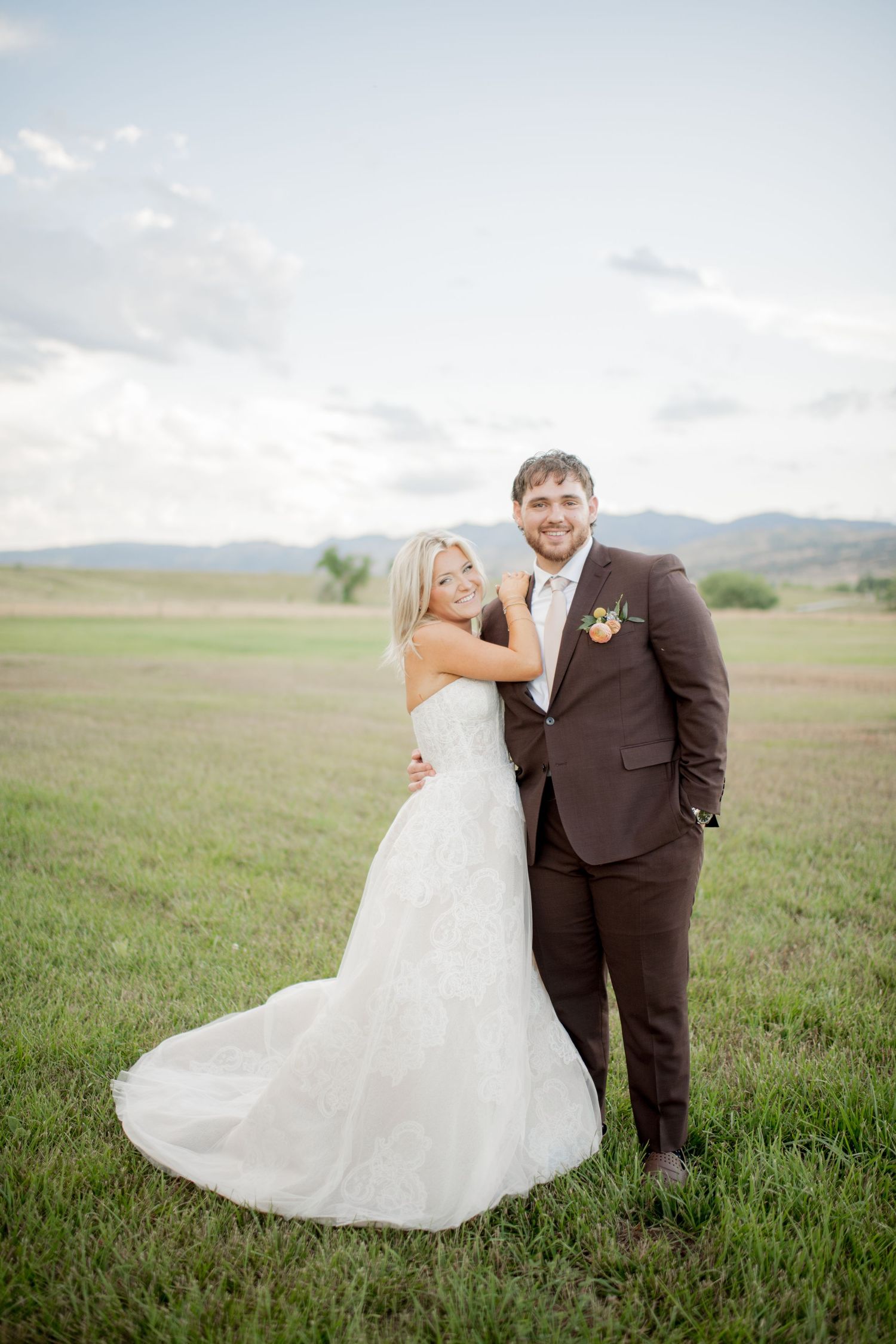 A couple in wedding attire stands together in a green meadow with rolling hills in the background.