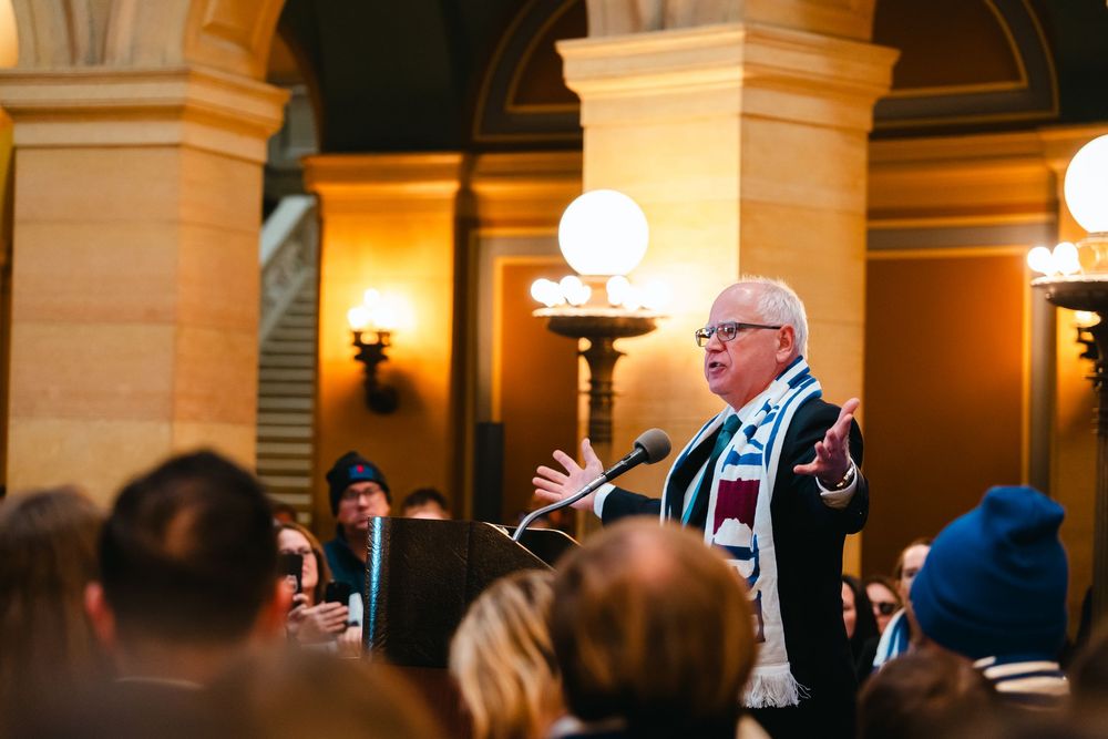 A person gestures while speaking at a formal indoor event with warm lighting and ornate architectural details in the background.