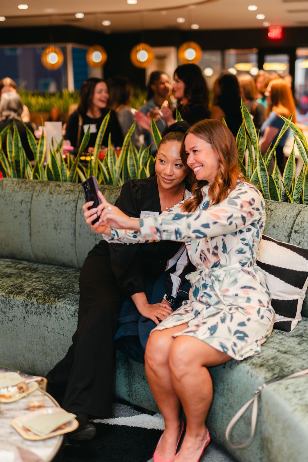 Two friends sharing a laugh while looking at a phone during a social gathering in an indoor venue with plants.