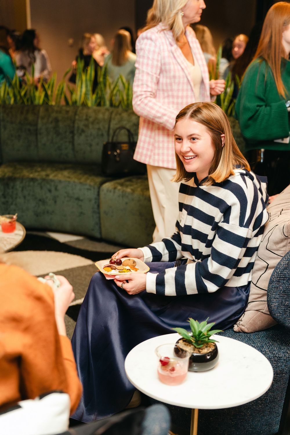 A person in a navy and white striped sweater sits on a couch enjoying a plated dessert at a social gathering.