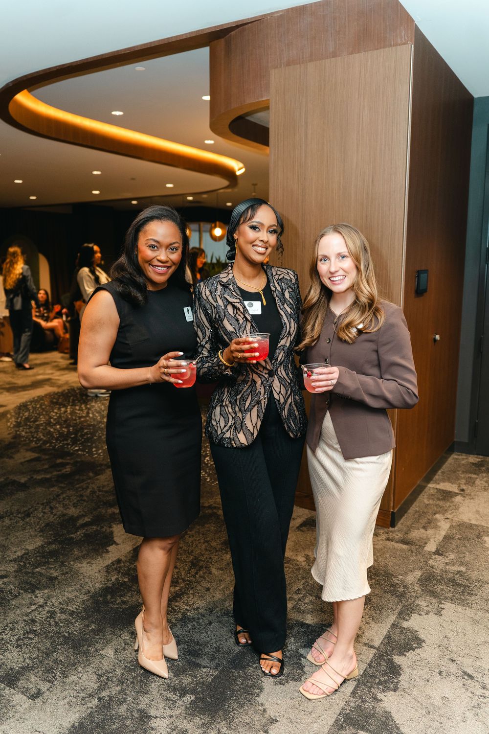 Three friends pose together at an indoor event holding red drinks in their hands.