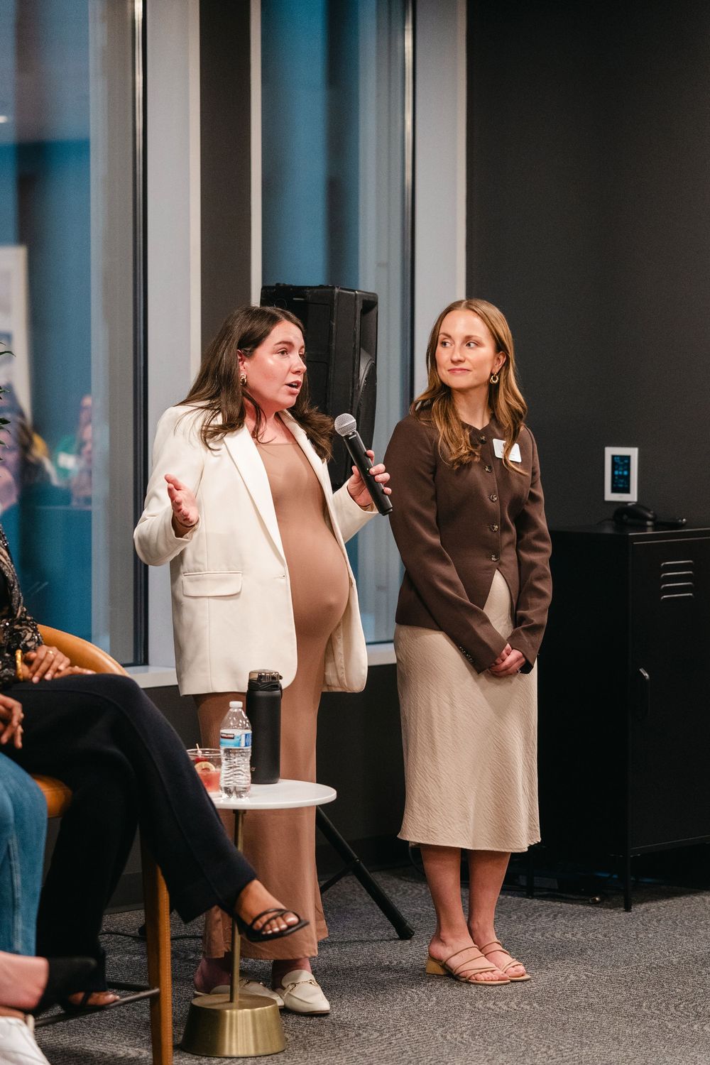 Two people stand at a speaking event, one wearing a white blazer and one in a brown coat with beige skirt.