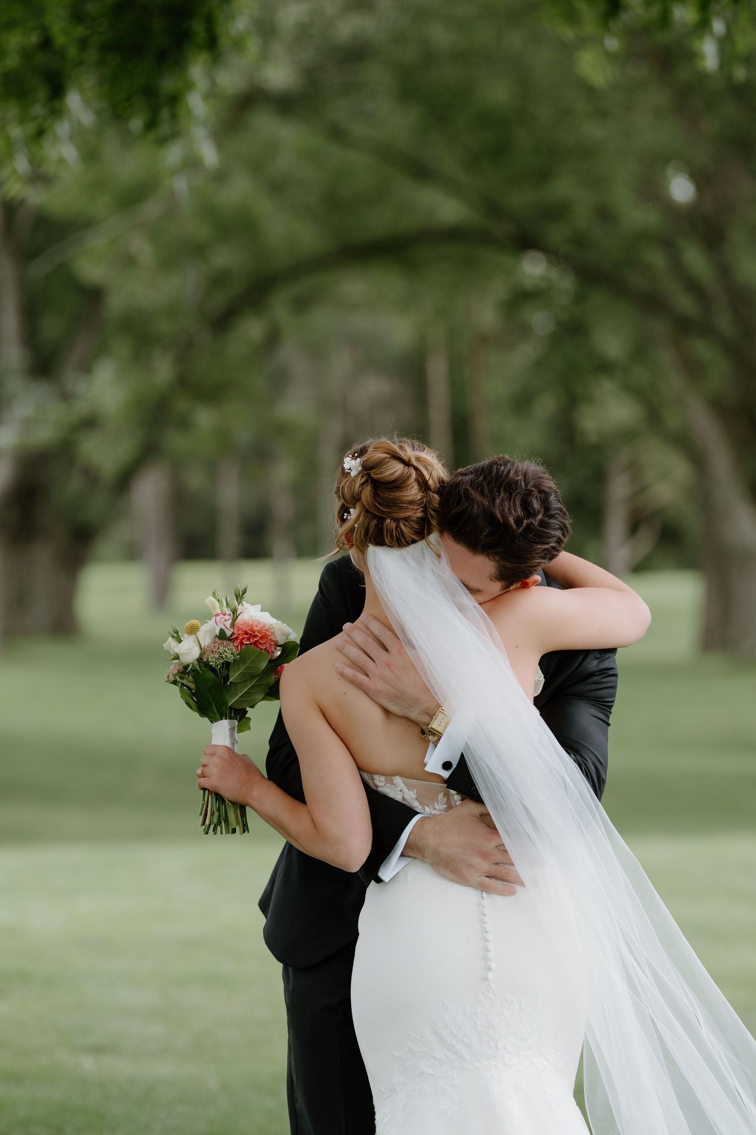 A bride in a fitted white wedding gown with lace details holds a peach and cream bouquet while walking on grass.