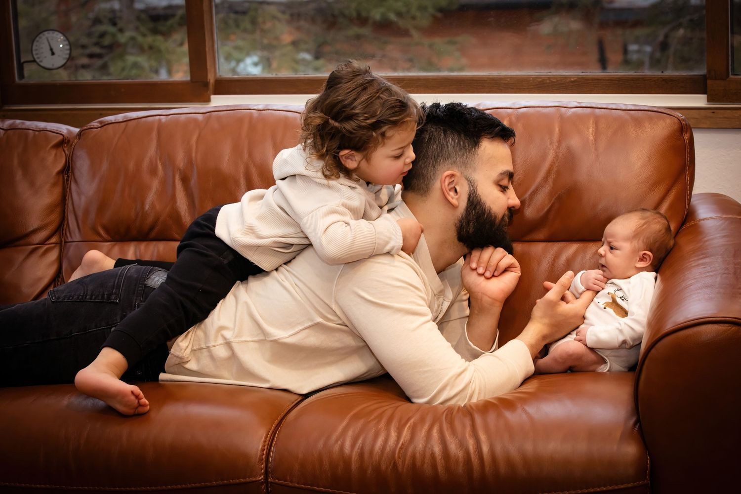 Family lying together on brown leather couch during cozy indoor photo session.