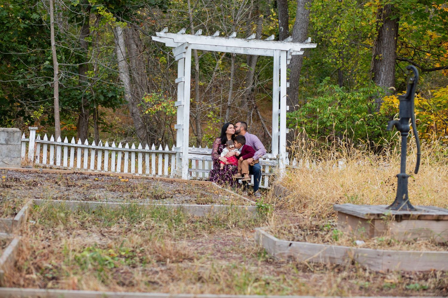 A white wooden arbor stands in a garden setting with autumn foliage and a picket fence in the background.