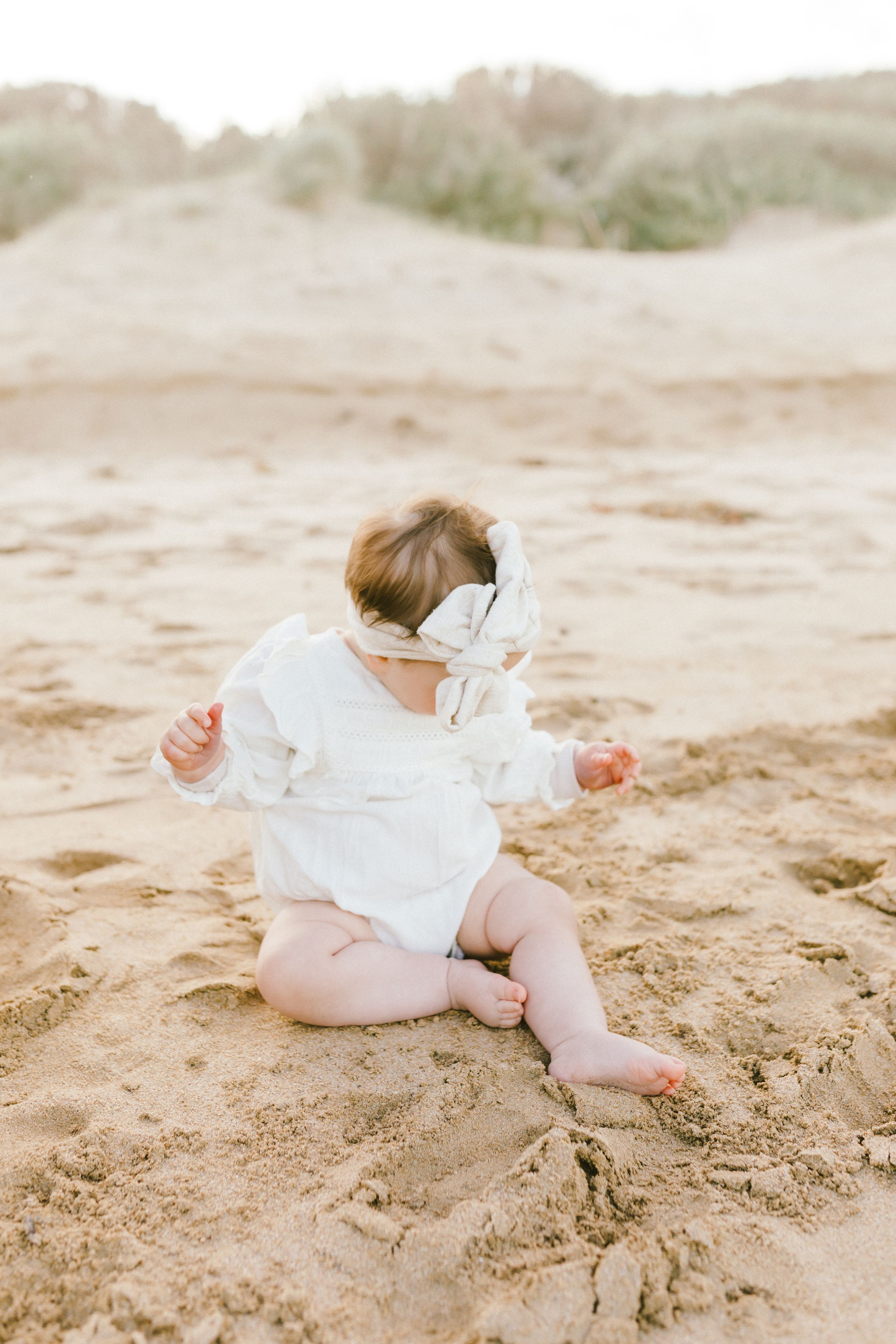 A baby in a white dress lies on soft sand at the beach during a photo session.