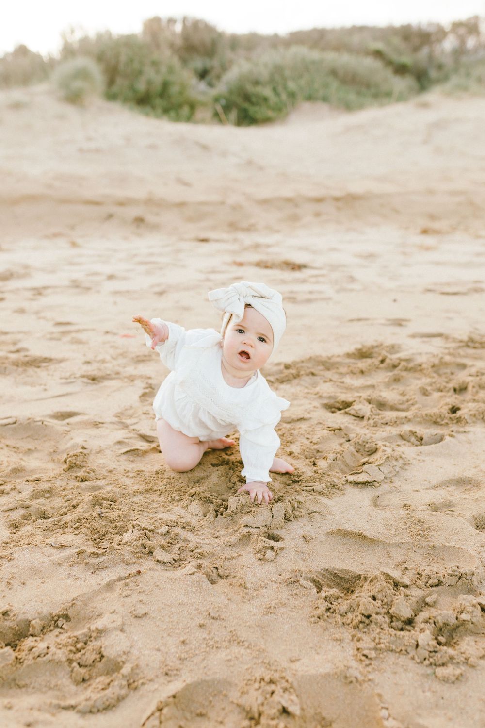 Baby in white clothing crawls and plays on sandy beach during outdoor photo session.