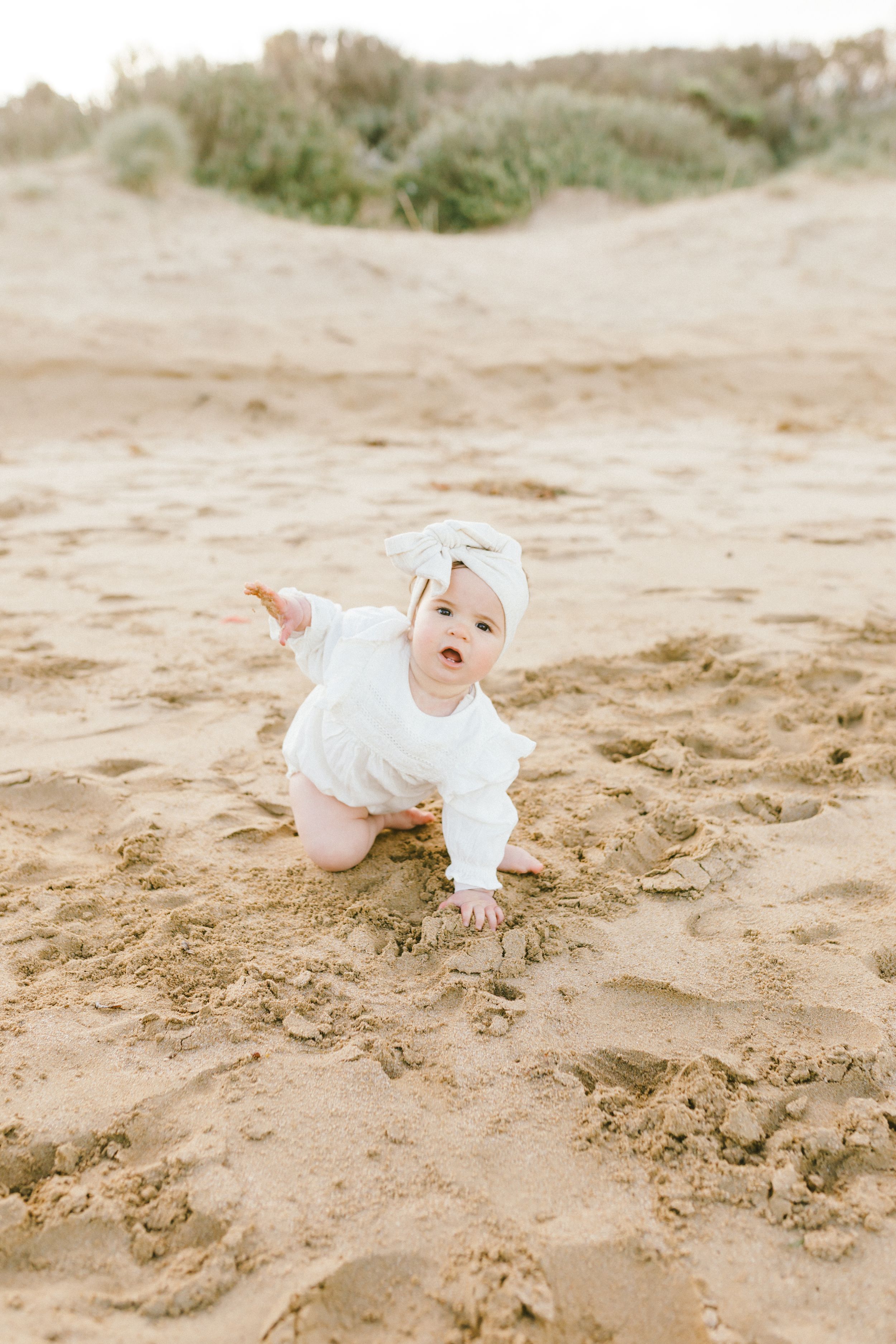 Baby in white clothing crawls and plays on sandy beach during outdoor photo session.